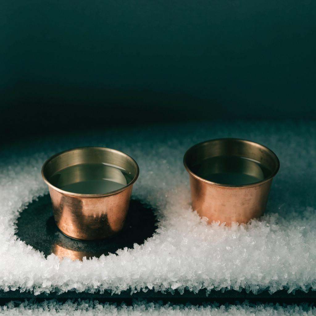 Two copper cups of water on a frosted surface, with a clear dark patch around the left cup, against a deep green background.