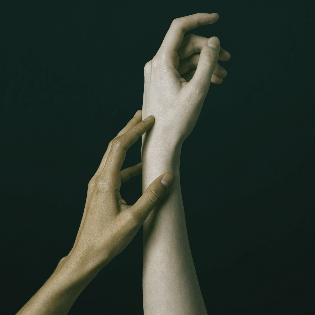 One hand touches the wrist and forearm of another raised hand with curved fingers, against a dark green background.